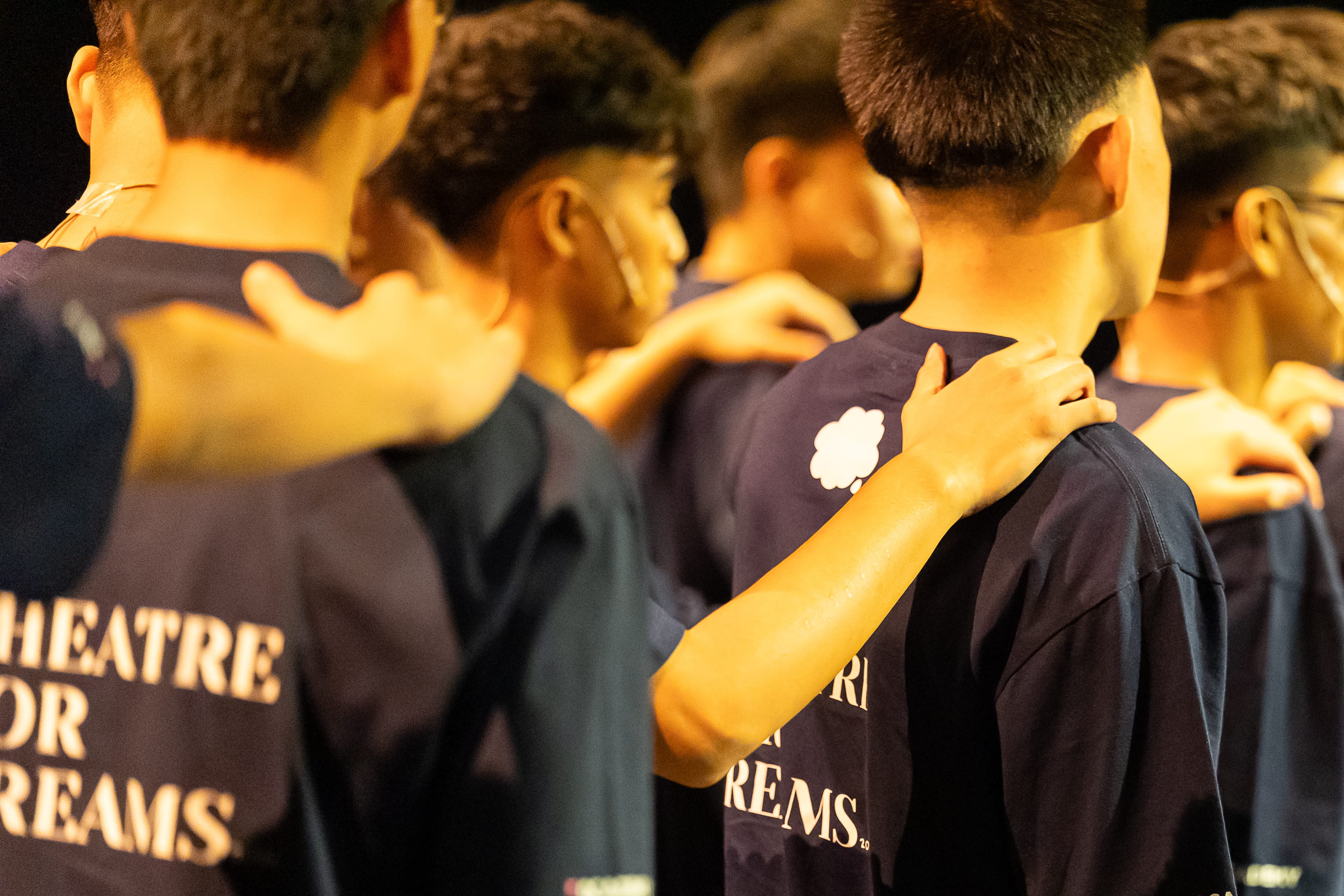 A group of young performers in matching 'Theatre for Dreams' t-shirts stand together with hands on each other's shoulders